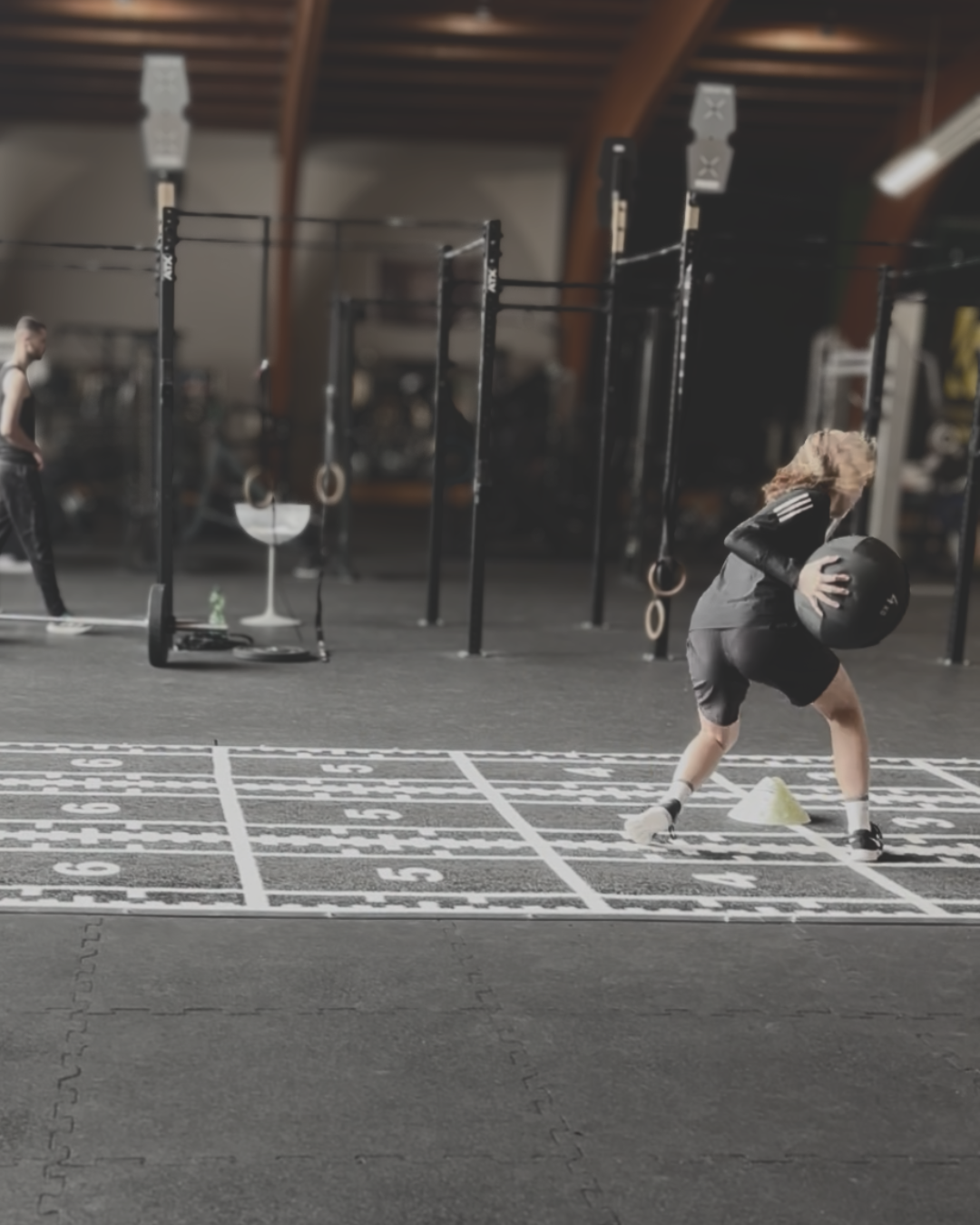 Athletin bei einer Agility-/Medizinball-Übung auf einer nummerierten Trainingsmatte in der Hi5Box, im Hintergrund Rig mit Ringen und weitere Trainierende