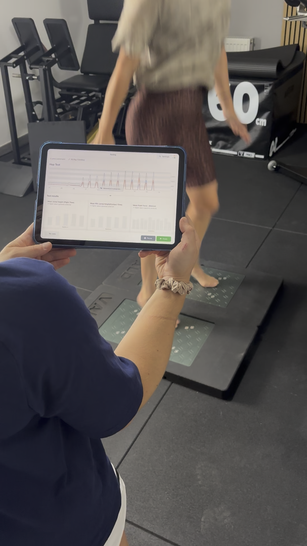Physiotherapeutin wertet live auf einem Tablet die Sprungmessdaten aus, im Hintergrund geht eine Person über die Kraft-/Sprungmessplatte mit sichtbarer 60cm-Markierung